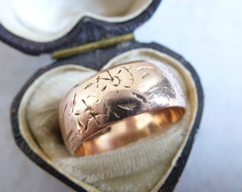 May include: A wide rose gold band ring featuring a series of small, hand-engraved starburst or floral motifs across its polished surface. The ring is positioned inside a dark brown, heart-shaped jewelry box with a cream-colored velvet interior.