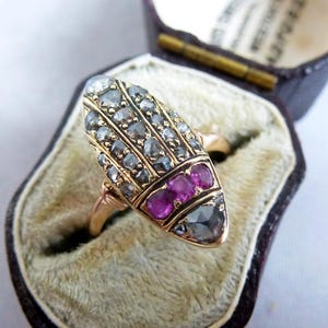 May include: A gold ring featuring an elongated, shield-shaped face set with multiple small, grey, faceted stones arranged in vertical rows, a horizontal band of three round, vibrant pink stones, and a single larger, triangular, grey faceted stone.