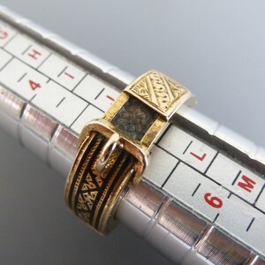 Puede incluir: Un anillo dorado dise&ntilde;ado para parecer un cintur&oacute;n con hebilla, con detalles en esmalte negro y una superficie texturizada. El anillo est&aacute; colocado en un mandril de anillo plateado, mostrando una medida de tama&ntilde;o entre 5 y 6 en la escala.