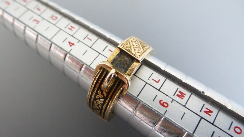 Puede incluir: Un anillo dorado dise&ntilde;ado para parecer un cintur&oacute;n con hebilla, con detalles en esmalte negro y una superficie texturizada. El anillo est&aacute; colocado en un mandril de anillo plateado, mostrando una medida de tama&ntilde;o entre 5 y 6 en la escala.