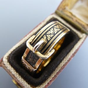 Puede incluir: Un anillo de oro dise&ntilde;ado para parecer un cintur&oacute;n con hebilla, con detalles de esmalte negro y patrones geom&eacute;tricos grabados a lo largo de la banda. El anillo se presenta dentro de una peque&ntilde;a caja rectangular con bisagras.