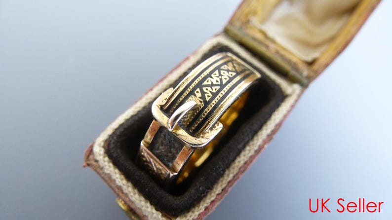 Puede incluir: Un anillo de oro dise&ntilde;ado para parecer un cintur&oacute;n con hebilla, con detalles de esmalte negro y patrones geom&eacute;tricos grabados a lo largo de la banda. El anillo se presenta dentro de una peque&ntilde;a caja rectangular con bisagras.