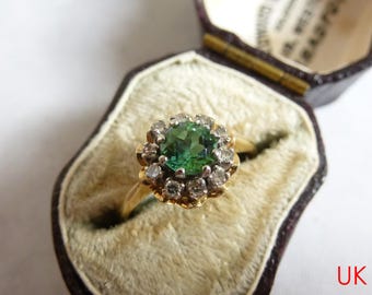 Puede incluir: Un anillo de oro con una piedra preciosa verde redonda y facetada en el centro, rodeada por un halo de pequeñas piedras claras de corte redondo. El anillo se presenta dentro de un joyero de color marrón oscuro forrado de terciopelo con el texto UK Seller.