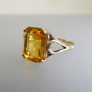May include: A gold ring with a large, clear, yellow gemstone set in an emerald cut. The ring has a delicate, openwork design.