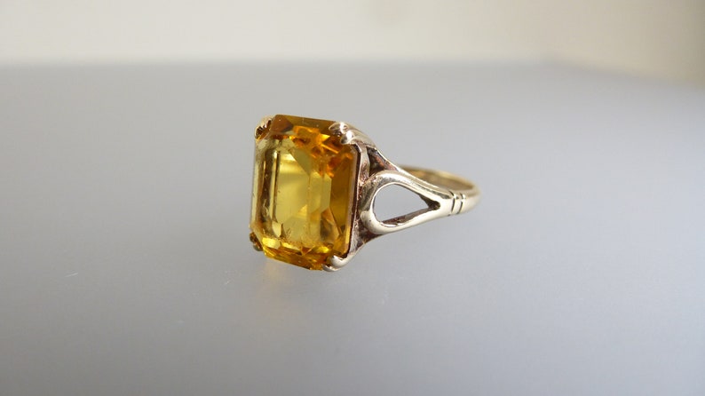 May include: A gold ring with a large, clear, yellow gemstone set in an emerald cut. The ring has a delicate, openwork design.