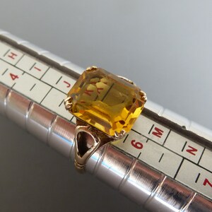 May include: A gold ring with a large, rectangular, yellow gemstone. The ring is sized on a ring sizer, showing a size 6.