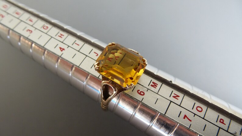 May include: A gold ring with a large, rectangular, yellow gemstone. The ring is sized on a ring sizer, showing a size 6.