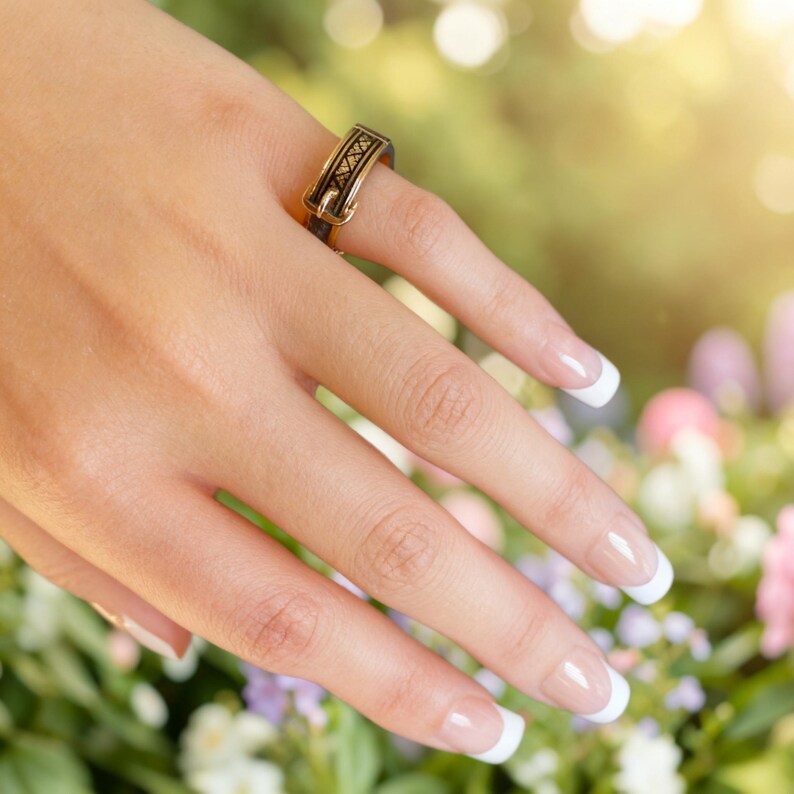 Puede incluir: Un anillo de tono dorado con un dise&ntilde;o de hebilla y una banda central negra. El anillo tiene un acabado met&aacute;lico pulido y un patr&oacute;n decorativo central que consiste en peque&ntilde;as formas geom&eacute;tricas repetitivas en la superficie exterior.