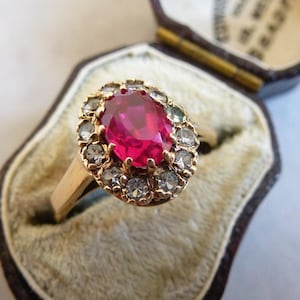 May include: A gold ring with a large oval ruby center stone, surrounded by smaller clear stones. The ring is displayed in a vintage jewelry box with a velvet interior. The ring's band is a simple, polished gold.