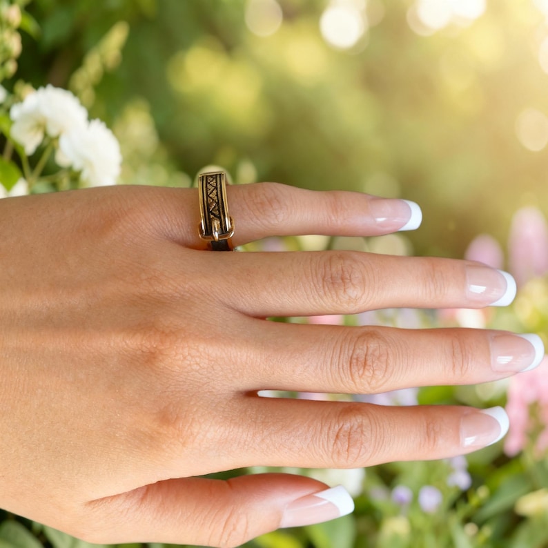 Puede incluir: Un anillo de tono dorado con un dise&ntilde;o similar a un cintur&oacute;n, que incluye una hebilla y detalles en esmalte negro. La banda presenta una superficie texturizada con un patr&oacute;n geom&eacute;trico repetitivo en el centro, como una correa en miniatura.