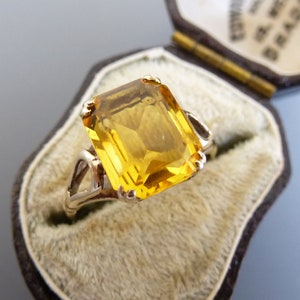 May include: A gold ring with a large, yellow, emerald-cut gemstone. The ring is in a velvet-lined box.
