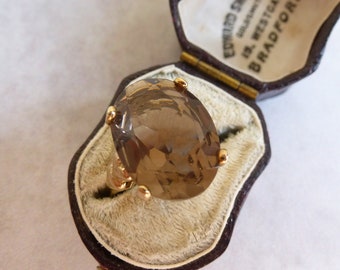 Puede incluir: Un anillo de oro con una gran piedra preciosa marrón translúcida de corte ovalado sujeta por cuatro garras. El anillo se muestra dentro de un joyero abierto forrado de terciopelo marrón oscuro con el texto Edward Smith, 19 Westgate, Bradford.