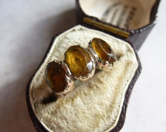 May include: A ring featuring three oval-shaped, translucent amber-coloured stones set in a silver-toned metal band. The ring is displayed inside a dark brown, hinged jewellery box with a cream-coloured, velvet-textured interior lining.