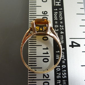 May include: A gold ring with a large, square-shaped, orange gemstone. The ring is measured against a ruler.