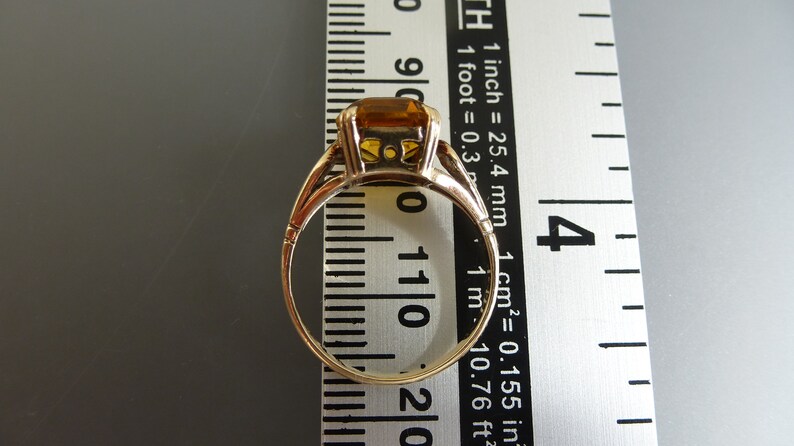 May include: A gold ring with a large, square-shaped, orange gemstone. The ring is measured against a ruler.