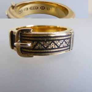 Puede incluir: Un anillo de oro dise&ntilde;ado para parecer un cintur&oacute;n con hebilla, con una incrustaci&oacute;n de esmalte negro con un patr&oacute;n geom&eacute;trico repetitivo. El interior de la banda muestra marcas claras, incluido el n&uacute;mero 625, que indica su composici&oacute;n.