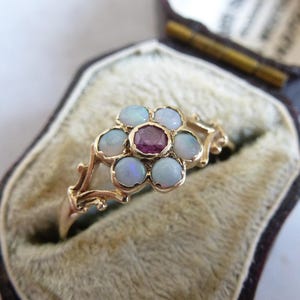 May include: A gold ring featuring a central round pink gemstone surrounded by six small, round, iridescent white opals arranged in a flower-like pattern. The band has a delicate, branching design and is presented in a dark brown velvet-lined box.