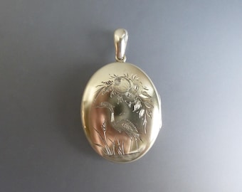 Puede incluir: Un colgante de relicario ovalado de metal dorado con un diseño grabado de un pájaro posado entre hierbas altas y elementos florales. El relicario tiene un acabado liso y pulido e incluye una pequeña anilla en la parte superior.