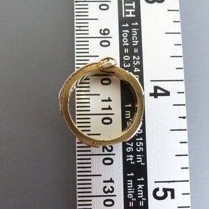 Puede incluir: Un anillo de metal en tono dorado con una banda texturizada y estampada colocado sobre una cinta m&eacute;trica blanca. El anillo tiene un di&aacute;metro interior de aproximadamente 1,9 cm, seg&uacute;n las marcas de la regla visibles a trav&eacute;s del centro.