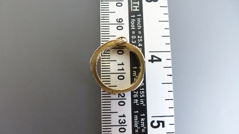 Puede incluir: Un anillo de metal en tono dorado con una banda texturizada y estampada colocado sobre una cinta m&eacute;trica blanca. El anillo tiene un di&aacute;metro interior de aproximadamente 1,9 cm, seg&uacute;n las marcas de la regla visibles a trav&eacute;s del centro.