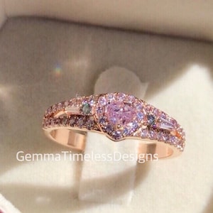 May include: A rose gold ring with a heart-shaped pink gemstone surrounded by smaller pink gemstones. The ring has a double band with additional smaller gemstones.