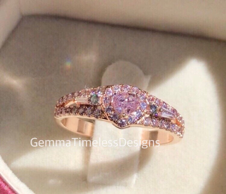 May include: A rose gold ring with a heart-shaped pink gemstone surrounded by smaller pink gemstones. The ring has a double band with additional smaller gemstones.