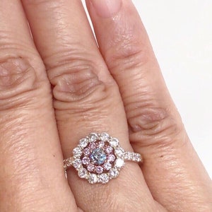 May include: A gold ring with a center stone surrounded by a halo of smaller diamonds. The center stone is a light blue diamond with a pink diamond halo.