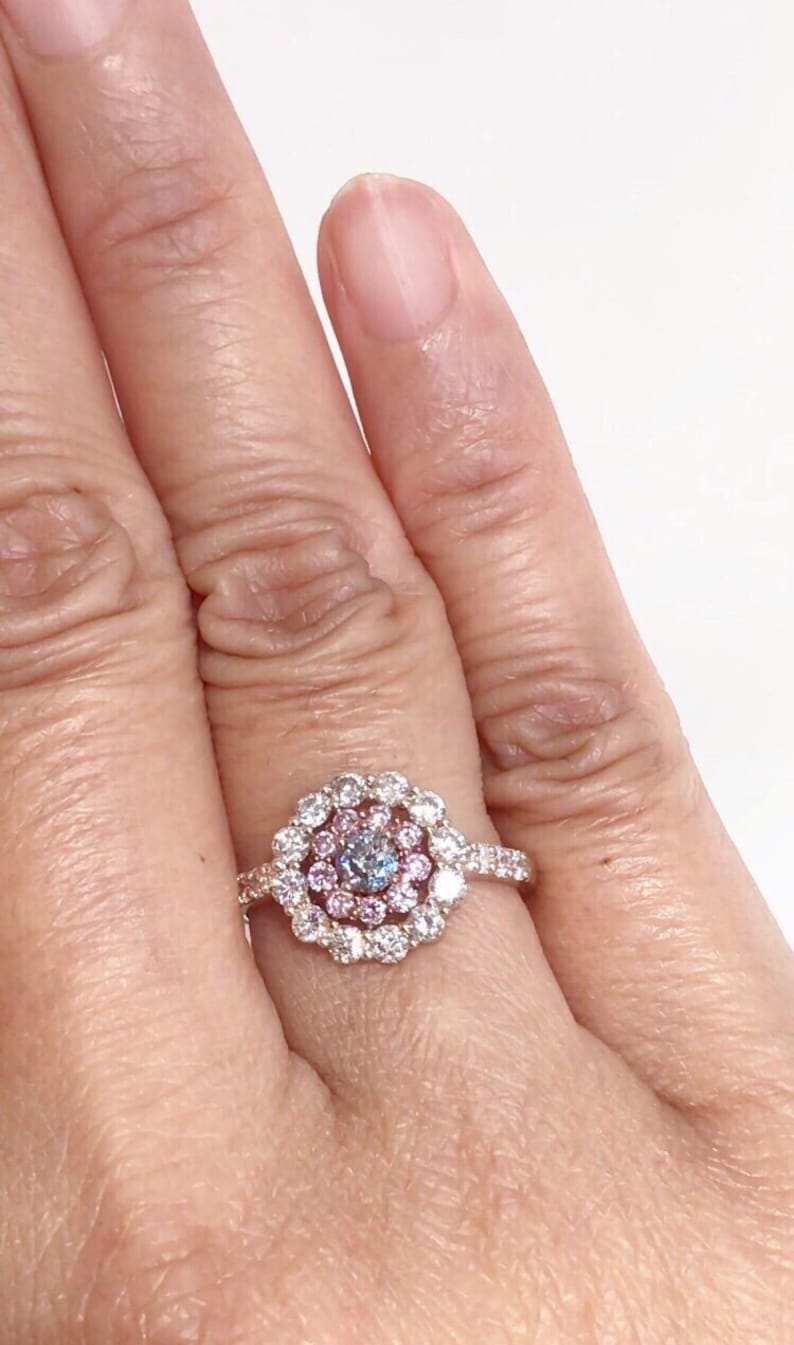 May include: A gold ring with a center stone surrounded by a halo of smaller diamonds. The center stone is a light blue diamond with a pink diamond halo.