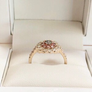 May include: A gold ring with a large center stone surrounded by smaller stones in a flower-like design. The center stone is a light gray color, and the surrounding stones are pink and white.