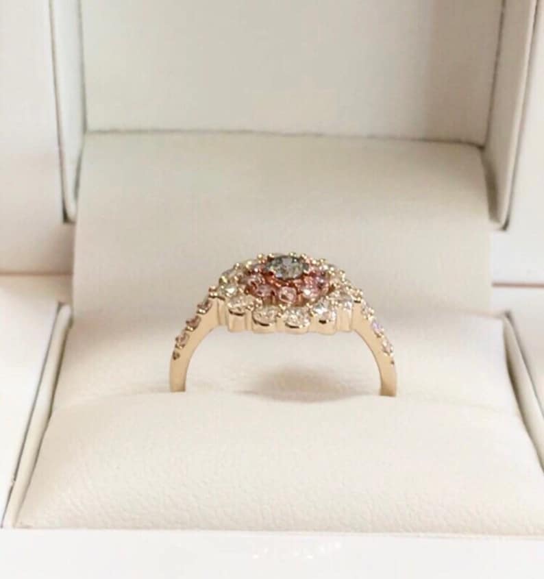 May include: A gold ring with a large center stone surrounded by smaller stones in a flower-like design. The center stone is a light gray color, and the surrounding stones are pink and white.