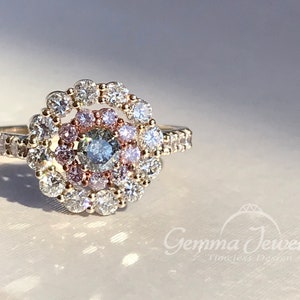 May include: A gold ring with a center stone surrounded by a halo of smaller diamonds. The halo is made up of two rows of diamonds, with the inner row being pink and the outer row being white. The ring is set on a white background.