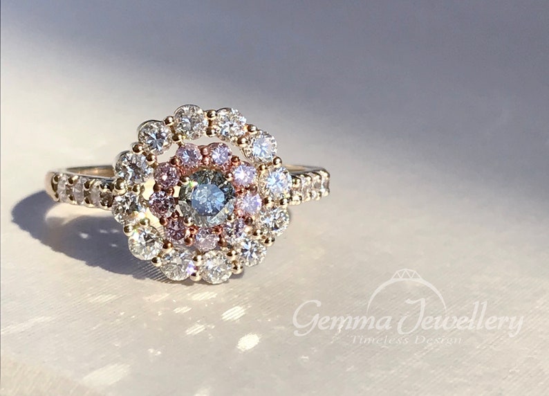 May include: A gold ring with a center stone surrounded by a halo of smaller diamonds. The halo is made up of two rows of diamonds, with the inner row being pink and the outer row being white. The ring is set on a white background.