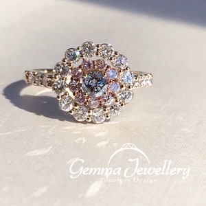 May include: A gold ring with a round center stone surrounded by a halo of smaller diamonds. The center stone is a larger diamond with a pink hue. The ring is set on a white background.