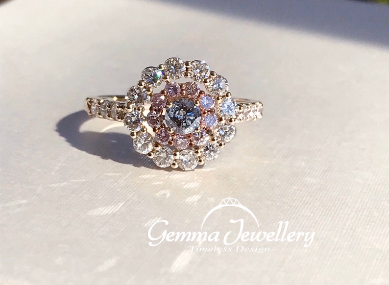 May include: A gold ring with a round center stone surrounded by a halo of smaller diamonds. The center stone is a larger diamond with a pink hue. The ring is set on a white background.