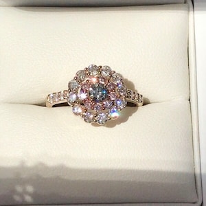May include: A gold ring with a large, round, multi-faceted center stone surrounded by a halo of smaller diamonds. The center stone is a light blue and gray color, and the halo is made up of white and pink diamonds.
