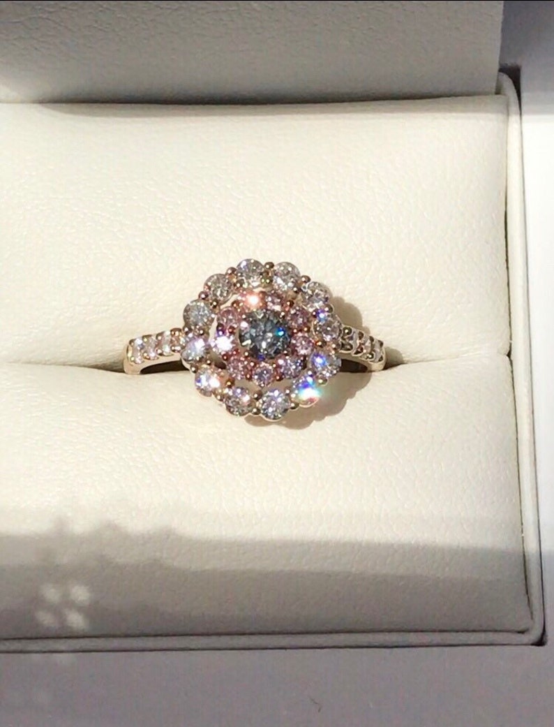 May include: A gold ring with a large, round, multi-faceted center stone surrounded by a halo of smaller diamonds. The center stone is a light blue and gray color, and the halo is made up of white and pink diamonds.