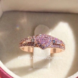 May include: A rose gold ring with a heart-shaped pink gemstone surrounded by smaller diamonds. The ring has a double band with additional diamonds.
