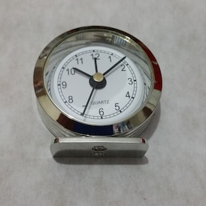 May include: A silver-toned analogue clock with a white face and black hands. The clock has a clear glass cover and a stand. The clock face has black numbers and the word "QUARTZ" printed on it.