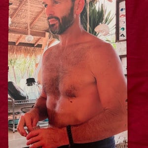 May include: Metal sign featuring a man with a beard, shirtless, in a tropical setting. The sign has a distressed look with visible wear and tear. The background includes a thatched roof and a framed display of images.