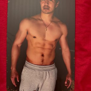 以下が含まれることがあります： グレーのスウェットパンツとネックレスを身に着けた、上半身裸の人物が描かれた金属製のサイン。サインは長方形で角が丸く、上部に2つの穴が開いており、吊り下げることができます。背景は暗いです。