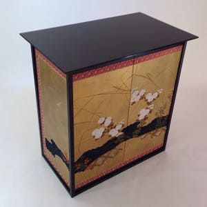 May include: A black wooden cabinet with two doors. The doors are decorated with gold leaf and a painted design of white flowers and black branches on a gold background.