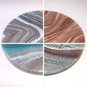 May include: A round, multi-colored resin serving tray with four different patterns. The tray has a white background with swirling patterns of gray, brown, teal, and pink.