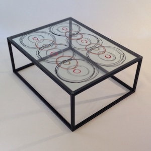 May include: A black metal framed glass coffee table with a geometric design of overlapping circles in black, red, and gold on the glass top.