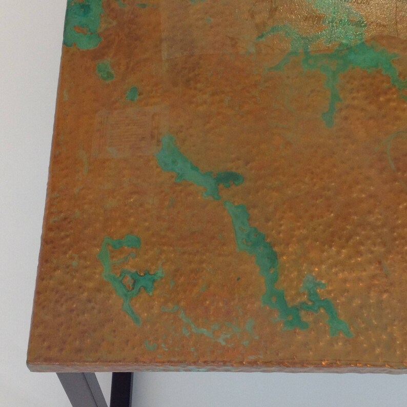 May include: A close-up of a copper table top with a hammered texture and green patina. The table top is made of metal and has a rustic look.