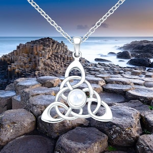 May include: A silver Celtic knot pendant necklace with a central, pale-coloured stone. The pendant hangs from a fine chain. The background shows a coastal scene with rock formations and the sea.