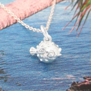 May include: A silver-colored necklace featuring a detailed pufferfish pendant. The pendant has a textured surface with small bumps and a smiling face. The necklace chain is delicate and silver, set against a blue and green background.