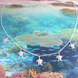 May include: A silver necklace featuring multiple small turtle pendants. The necklace has a delicate chain and the turtle charms are evenly spaced. The background shows an underwater scene with coral and fish.