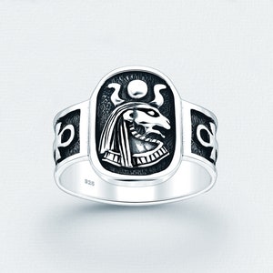 May include: A silver ring with an oval centerpiece featuring an Egyptian deity with a sun disc. The ring has an intricate design with ankh symbols on the sides. The ring is made of 925 silver.