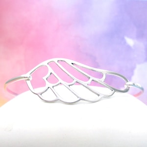 May include: A silver-colored bangle bracelet featuring a stylized wing design. The wing is open and detailed with a delicate, openwork pattern. The bracelet is set against a soft, colorful background.