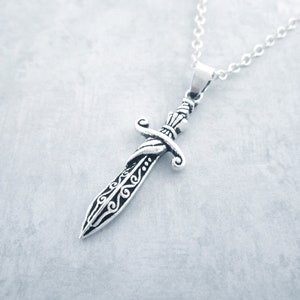 May include: A silver-colored pendant necklace featuring a detailed dagger design. The dagger has an ornate handle and blade with decorative patterns. The necklace chain is also silver, creating a cohesive and elegant accessory. The pendant is approximately 2 inches long.